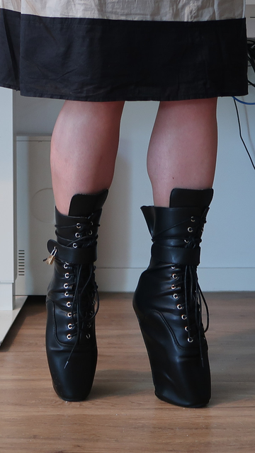 A knee down picture of me wearing kinky boots. Those are very high wedge heels, keeping the foot in en pointe position. The boots are reaching the mid-calf. They are laced. On each boot there is a wide strap at the ankle. On one boot it is visible, the strap is locked with a padlock. It is impossible to take them without unlocking the padlock, or damaging the boots. Practically, the person wearing those boots is at mercy of whoever keeps the keys to padlocks. Maybe some willing lock picker would give some relief, but what they will expect in exchange?