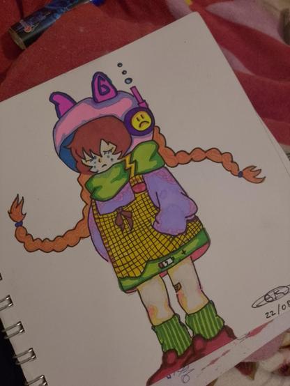 Un dibujo de un personaje estilo cartoon de cuerpo completo, con una expresión melancólica y lágrimas en los ojos. Tiene cabello castaño rojizo con flequillo y dos largas trenzas que se extienden a los lados. Viste un gorro grande con orejas de gato, en tonos rosa y azul, y lleva auriculares morados con un emoji de cara triste en uno de los audífonos. De su gorro emerge un tubo estilo snorkel con pequeñas burbujas flotando. Lleva una bufanda verde alrededor del cuello y un suéter o chaqueta holgada de color lila. Debajo, un vestido o peto amarillo a cuadros con un lazo marrón en el pecho. En el dobladillo inferior del vestido, se ve un icono de batería con poca carga, sugiriendo fatiga. Sus piernas son de tono claro, con calcetines a rayas verdes y zapatos rojos. El dibujo, realizado con colores brillantes, está en una hoja de cuaderno con la espiral visible a la izquierda.