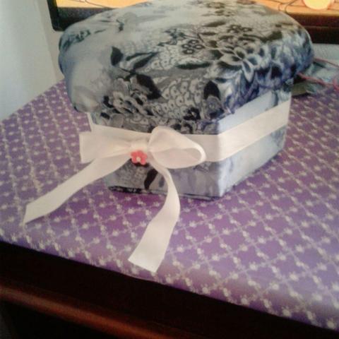 Una caja decorativa de tela con un patrón floral y ornamental en tonos de azul, gris y blanco. La caja es poligonal y tiene una tapa ligeramente abombada. Está adornada con una cinta de satén blanca que la envuelve, anudada en un lazo prominente en la parte frontal. En el centro del lazo hay una pequeña flor decorativa de color rosa pálido. La caja descansa sobre una superficie de tela de color lila o morado claro, que presenta un patrón de cuadrícula de pequeños puntos blancos. El fondo es borroso, mostrando un mueble oscuro de madera en la parte inferior.