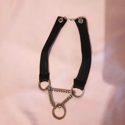 Una gargantilla o collar de estilo alternativo, compuesto por dos tiras de material negro (posiblemente cuero sintético) que se unen en la parte superior con un pequeño broche metálico plateado. En la parte inferior, cada tira se conecta a un anillo metálico plateado. De estos dos anillos cuelgan cadenas plateadas que forman un triángulo, uniéndose en un anillo metálico más grande en la parte inferior central. El accesorio está apoyado sobre una superficie de tela rosa pálido, ligeramente arrugada.
