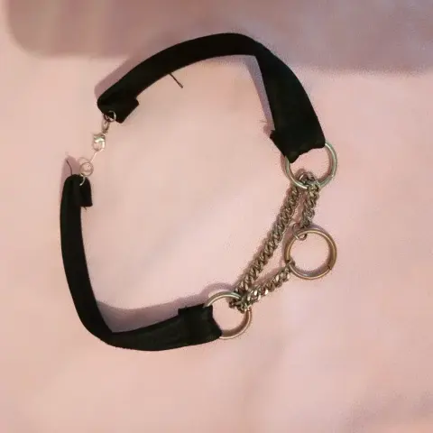 Una gargantilla o collar tipo choker de color negro y metal plateado, dispuesto sobre una superficie lisa de color rosa claro. La gargantilla se compone de una banda ancha de tela negra, que parece terciopelo o una cinta, con un cierre de langosta plateado en un extremo. De la banda cuelgan tres anillos metálicos plateados, interconectados por dos cadenas finas también de metal plateado, creando un efecto de capas o cascada en la parte frontal.