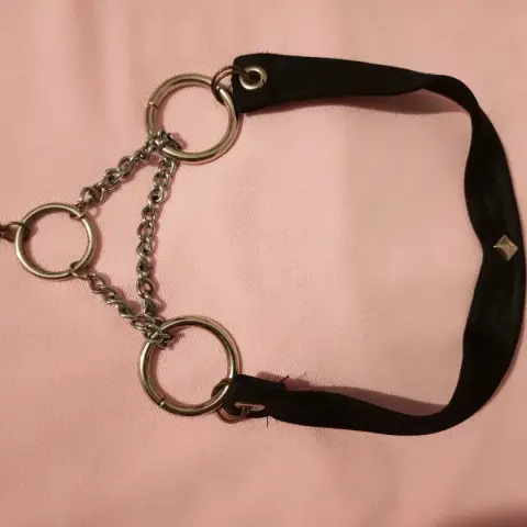 Una gargantilla o accesorio de estilo gótico/punk. Consiste en una banda ancha de material textil negro, curvada en forma de U, con un remache metálico plateado en forma de pirámide en su lado derecho. En cada extremo de la banda, un gran anillo metálico plateado está sujeto por un ojal. Desde estos dos grandes anillos, dos cadenas finas de eslabones plateados se extienden hacia el centro, donde se unen a un anillo plateado más pequeño. Este anillo más pequeño se conecta con otra cadena a un cuarto anillo plateado grande en el extremo izquierdo. El accesorio está sobre una superficie de tela rosa claro.