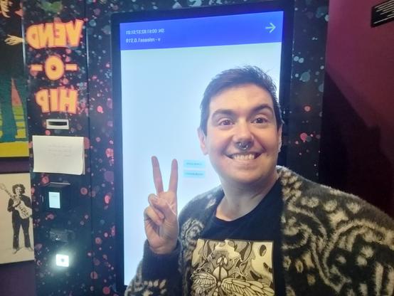 I'm taking a beaming selfie in front of a hippie vending machine (the Vend-O-Hip) which is the kind with an enormous screen on it. It has a blue and white screen on it with some developer options.