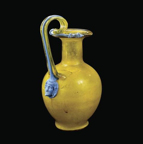 The picture shows a yellow pitcher. The handle is decorated with a blue glass thread that terminates in an appliqué with a woman's head.