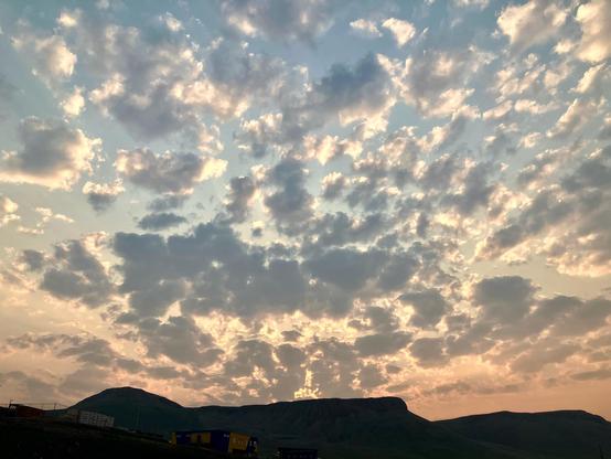 Dark hills that ring town. Above them are an explosion of clouds, blue, grey, and white. The sky intermingling the clouds is pink, orange, and yellow