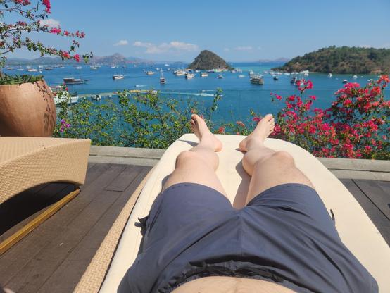 Lying on a beach bed, wearing shorts. Beyond patio, ocean, boats, many islands. Blue sky, clouds only in the distance.