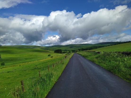 Is single lane tarmac road leading through rolling hilly green countryside under blue sky with 5/10 cloud cover. You cannot hear anything.