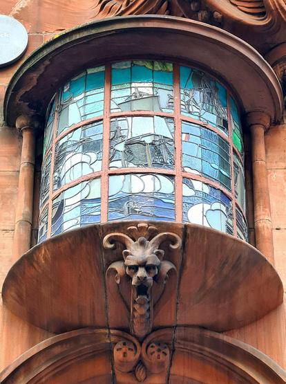 A curved stained glass window featuring ships on a stormy sea, with the mask of a horned creature below it.