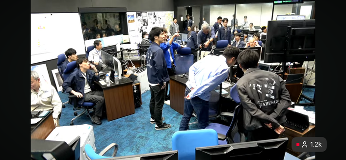 Scene from the control room during the flyby observations of asteroid Torifune by Hayabusa2. Several team members are wearing jackets with an image of the flyby on the back.