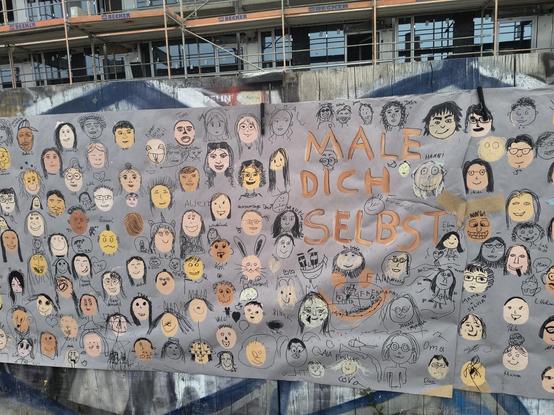 Ein Plakat "Male dich selbst". Darauf sind viele Ovale in unterschiedlichen Hauttönen gemalt. Menschen gehen mit Edding daraus Gesicht gemalt