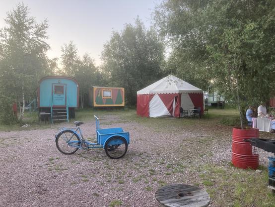 två cirkusvagnar varav en är fakirens. även ett mindre partytält (oklart om det också var ett hotellrum) samt en blå snygg gammal trehjulig lastcykel.
grusig plan, grönskande omgivning.