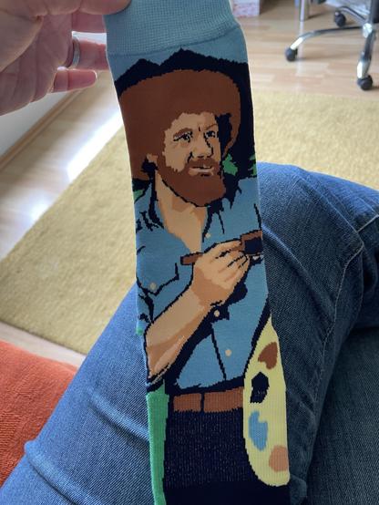 Eine bunte Socke mit einer Grafik von Bob Ross. Mit lockigem Haar und Bart, er hält eine Farbpalette.