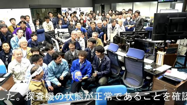 screenshot from the live stream. In the Hayabusa2 control room,  the team are gathered before the camera, all beaming. Front and centre in a bright blue jacket is a young man holding a blue spherical Daruma doll.