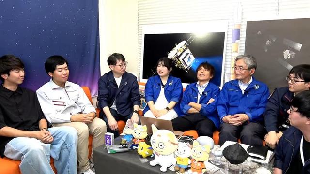 screenshot from the live stream, showing the livestream narrators, now with team leader Mimasu Yuya, chatting around a small table. Yuya looks exhausted!