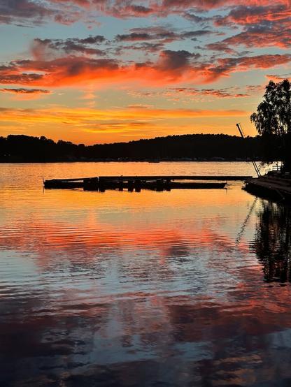 a sunset scene over a body of water. orange-lit clouds reflect in the water, and the silhouette of a pier breaks up the golden glow