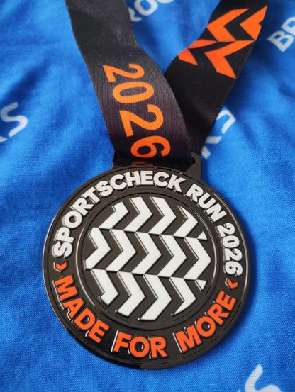 A medal on a blue background.
The medal says "SportScheck Run 2026. Made for more". It's a black medal with a white zig-zag pattern in the middle. The writing is in white and orange.
The blue background has the name "Brooks" in  white in a repeating pattern.