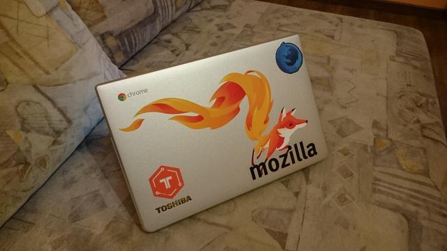 A Toshiba Chromebook on a couch, the lid is open with the cover facing the camera. Top left corner is the embedded "Chrome" chromebook logo, top right corner has a circular blue Firefox sticker representing the "Developer Edition" of the browser. Bottom left corner above the Toshiba mark is a hexagonal red "Tessel" logo sticker for the microcontroller platform of late 2010s. But the prime eye catcher is the fox in the bottom right corner, above the "mozilla" word-mark, with her blazing, swirling tail reaching all the way across the cover into the top left corner.
