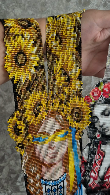 The hand holds two long. intricate Ukrainian gerdans (beaded jewelry), each decorated with a beaded portrait of a Ukrainian woman. The left one, with vellow and blue hues. features sunflowers and a woman wearing patriotic glasses. The right one, in white, black, and red, shows a woman in traditional dress. Both have beaded fringe.