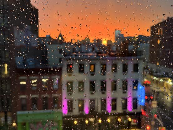 A rain-drenched window offers a blurred view of city buildings illuminated by a vibrant sunset, with clouds painted in shades of orange and purple. Water droplets create a textured foreground, enhancing the scene's atmospheric feel.