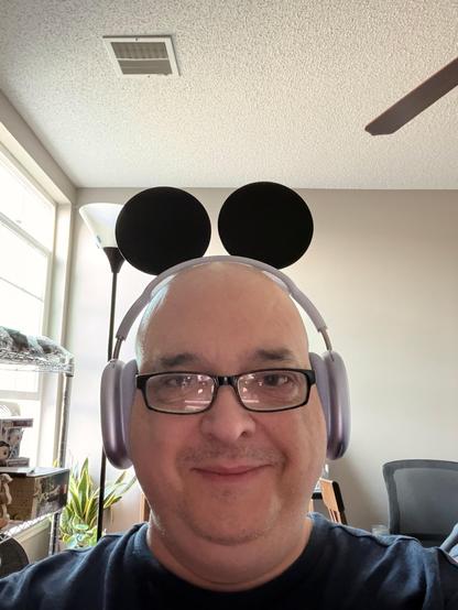 Smiling, round faced man with glasses and headphones on looks at the camera. The prominent feature of the photo are two famous rodent shaped ears attached to the headphones. The man is smiling while thinking of the whimsy of it all.