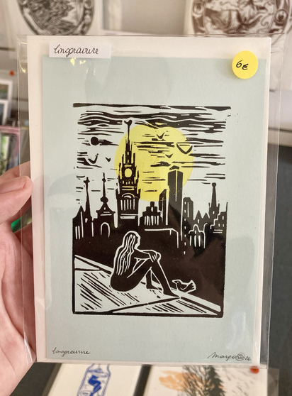 A A6, postcard sized, linocut print in two colours. Printed in black is a skyline of the city of Antwerp. In the front, a woman is sitting on the roof looking over the city with a bird next to her. In the background, behind the cathedral, is a yellow sun.