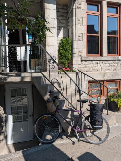 A photo of a basic bike with an overstuffed pannier on the rear and a canvas postbag style purse tied to the handlebars, in front of a stone building with curving stairs up to an open door and stairs down to a closed door.