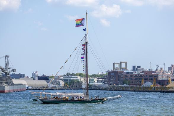 A small, single-masted sailing vessel on a river. Behind it is a dockyard and some buildings. The vessel’s sails are lowered, and it flies a large Progress Pride flag.
