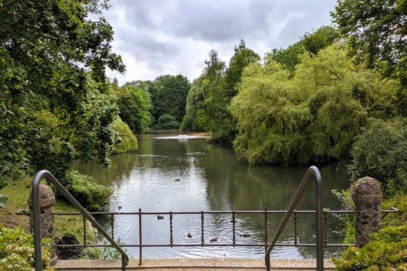 Blick vom oberen Ende einer Treppe auf eine Metallbrüstung, dahinter ein kleiner See mit einer Art Wassersprudler in der Mitte. Rundherum Bäume, der Himmel ist bewölkt. Ein paar Enten paddeln auf dem Wasser.