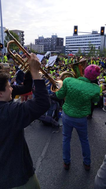 Musiker einer Band im Vordergrund, im Hintergrund eine Menge von Menschen mit gelben Warnwesten in einem Stadtgebiet mit Häusern
