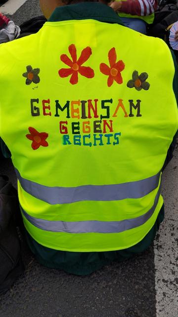 gelbe Warnweste mit dem Schriftzug "gemeinsam gegen Rechts" in bunten Buchstaben geschrieben; dazu einige Blüten