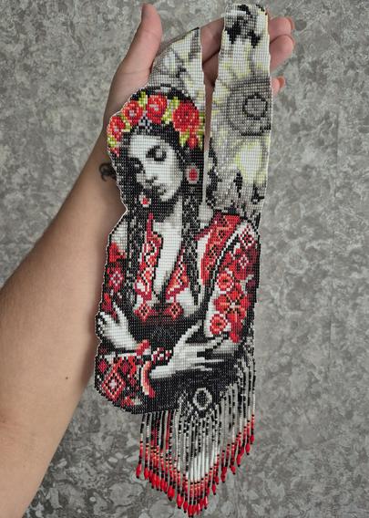 A traditional Ukrainian beaded ornament (herdan) held in the hand. The product features a detailed portrait of a girl in authentic attire: a red-and-black embroidered shirt and a bright wreath of flowers. The lower part of the ornament is decorated with a beaded fringe with red accents at the ends. The photo was taken against a gray textured wall.

,