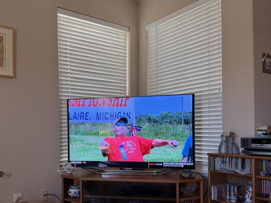 A photo of a tv in a corner of a room, showing a man standing in the field of grass, leaning back, with his cheeks puffed and his lips puckered