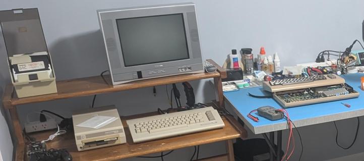 On the left is a wooden desk with a C64C, CRT TV, 1541 floppy drive, and a box full of floppies. On the right is a workbench with a breadbin C64 opened on a static mat. There is additionally a MiSTer set up on the desk.