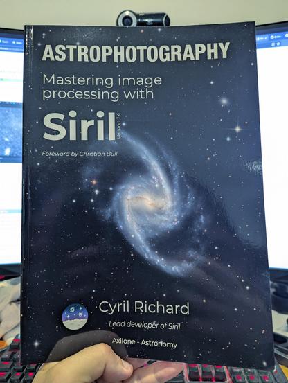 Photo of a person holding up a glossy A4 book with a picture of a galaxy on the cover, titled "Astrophotography: Mastering Image Processing with Siril".