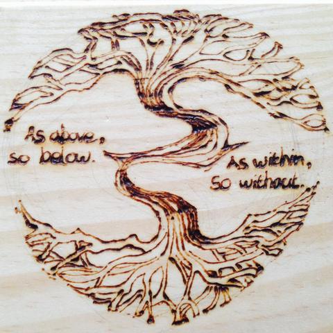 A simple drawing using pyrography (wood burning) of a tree with symmetrical roots and branches. Words around it read, “As above, so below. As within, so without.”