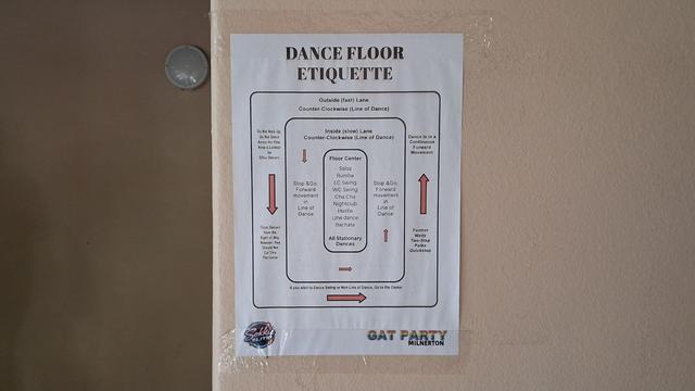A poster stuck on a cream-painted institutional-looking wall with a round light in the background. 

The title of the poster in big black letters is "DANCE FLOOR ETIQUETTE" and it shows a floor plan with concentric rounded rectangles, each with labels and red arrows indicating a counter-clockwise movement. 

 The outer rectangle is titled "Outside (fast) Lane Counter Clockwise (Line of Dance)" and  includes the notes "Do not back up, do not dance across the flow, keep a lookout for other dancers" and "These dancers have the right of way, however, they should not cut through the center", "Dance is in a continuous forward movement", "If you wish to Dance Swing or Non-Line of Dance, Go to Center" and "Foxtrot, Waltz, Two-step, polka, quickstep". 
The next rectangle inside the first is titled "Inside (slow) Lane Counter Clockwise (Line of Dance)" and includes the note "Stop & Go, Forward movement in line of dance". 
The central rectangle is labeled "Floor Center" and includes a list of dances "Salsa, Rumba, EC Swing, WC Swing, Cha cha, Nightclub, Hustle, Line Dance, Bachata All Stationary Dances"
At the bottom of the poster in big rainbow coloured letters it says "GAT PARTY" and in black letters "Milnerton" as well as a logo that says "Sokkie Elite"