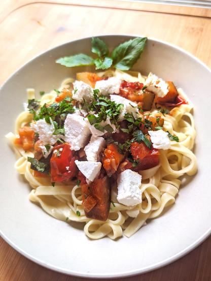 Ein tiefer Teller mit Tagliatelle, darauf buntes Ratatouille aus Zucchini, Paprika und Tomaten. Darüber grob zerbröselter Feta, gehackte Petersilie und frische Basilikumblätter als Garnitur. Das Gericht steht auf einer Holzarbeitsplatte und ist von warmem Tageslicht beleuchtet.