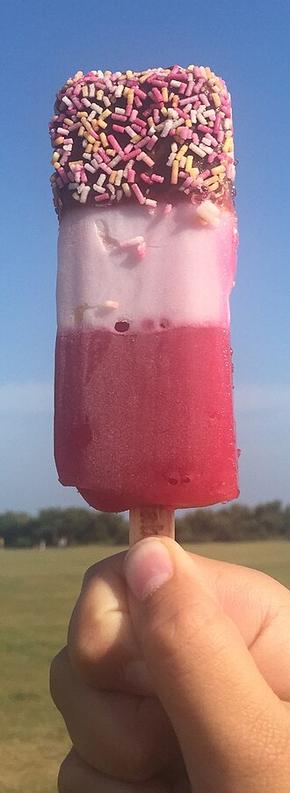 A hand holds a multicoloured ice lolly against a clear blue sky and a blurred grassy field. The ice lolly consists of a dark chocolate top layer coated in pink, yellow and white sprinkles, followed by a pale pink middle section and a deep red base.