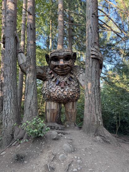 Wooden statue of a large troll between some trees. His arms are stretched out to hold the trees on either side of him.