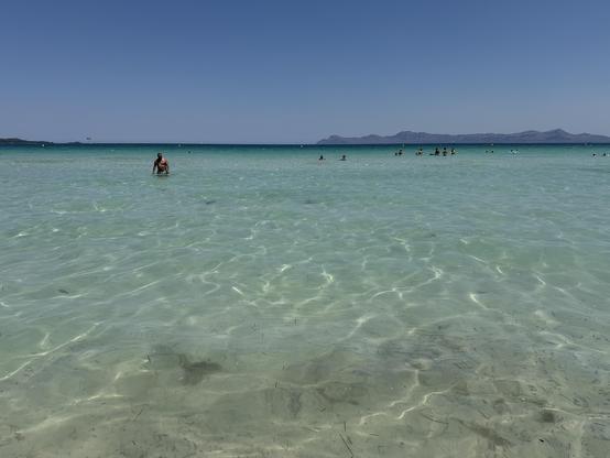 A photo of a sea taken from the shallows. The sea is clear and gradient to the nicest aquamarine. The sky is blue. There are mountains on the horizon to the right.