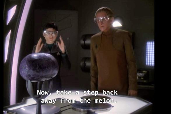 DS9 scene. Ezri Dax and Odo (🎶 Bucket's of fun🎶) are standing next to a table. Both are wearing clear safety glasses, and both are human looking though one has a slug worm thingy inside of her and the other is a shapeshifter guy with v smooth features. On the table is a round purple object being held up by some kind of holder-upper type thing. Closed caption reads, "Now, take a step back from the melon."