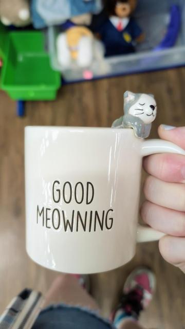 A large, creme-colored mug, with a ceramic cat hanging out of it. The mug says "Good Meowning" on the sides.
