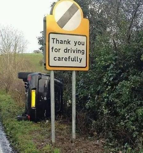 Uncredited meme with a photo of a government-installed traffic safety sign seemingly placed along a rural country road, and the public sign says, "Thank you for driving carefully". Behind this sign we see a black SUV lying on its right side whilst its occupants (if any) are not visible.