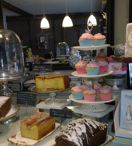 A display of various baked goods in a café, featuring a lemon drizzle cake, a pear and ginger cake, pink frosted cupcakes on a stand, and a banana loaf, all presented with elegant labels. The background includes a mirror and decorative items.
Eine Auslage verschiedener Backwaren in einem Café, darunter ein Zitronenkuchen, ein Birnen-Ingwer-Kuchen, Cupcakes mit rosa Zuckerguss auf einem Ständer und ein Bananenbrot, die alle mit eleganten Etiketten versehen sind. Im Hintergrund sind ein Spiegel und Dekorationsgegenstände zu sehen.