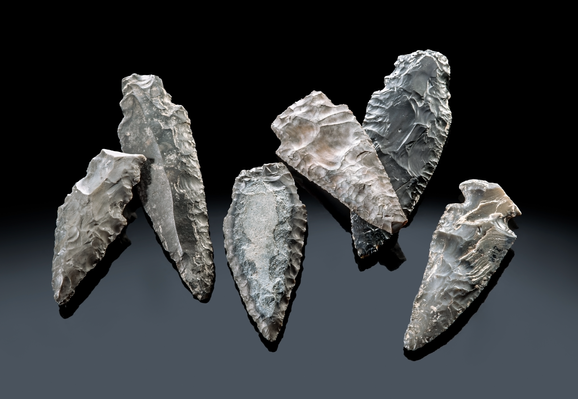 Six neolithic flint dagger blades from Lake Constance showing skilled pressure flaking and varying colors. They are displayed against a dark background.