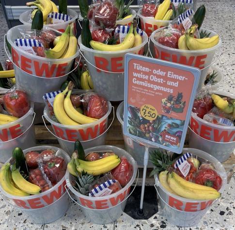 Auf und vor einer Palette sind Zehn-Liter-Kunststoffeimer mit der Aufschrift REWE mit Obst und Gemüse gestapelt. Ein Schild preist “Tüten- oder Eimerweise Obst- und Gemüsegenuss für nur 7,50 Euro” an.