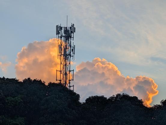 A photo of a cell tower against a blue sky, setting sun backlighting cumulus clouds in warm colours. A low canopy in silhouette in the foreground