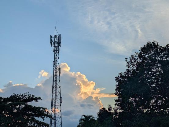 A photo of a cell tower against a blue sky, setting sun backlighting cumulus clouds in warm colours. Trees in silhouette in the foreground