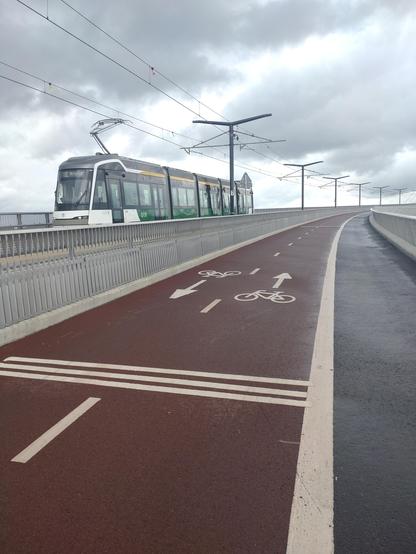 Detailbild das den Radweg zeigt. Der Radweg hat rot eingefärbten Asphalt unf isr mit einer Mittelmarlierung in zwei Fahrtrichtungen geteilt. Auf den Straßenbahngleisen fährt in diesem Moment eine Bahn vorbei.