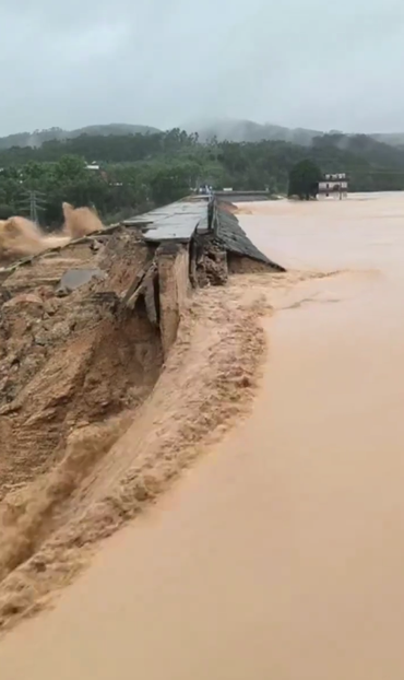 Still frame from a video showing a tall, collapsed dam through which massive stream of brown water if rushing